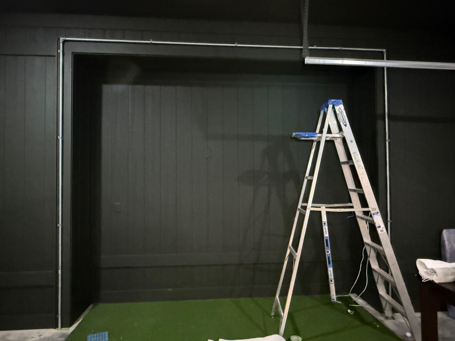 Dedicated golf simulator room with dark walls and turf during setup